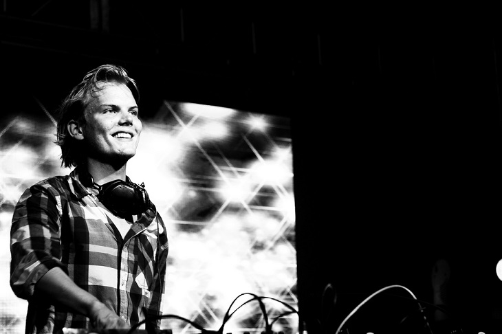 Avicii står bakom ett mixerbord. Han har en rutig skjorta och halvlångt hår som är struket bakåt. Han tittar ut mot publiken och ler.